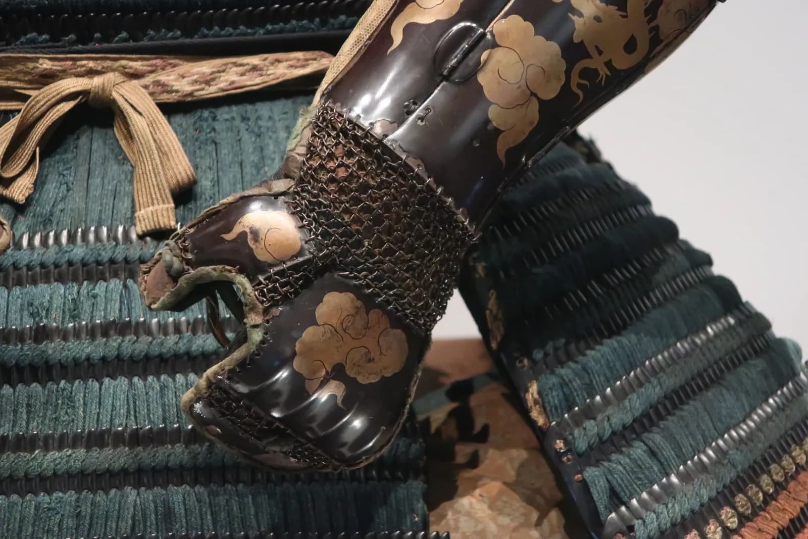 A close-up view of the intricate details of traditional samurai armor, featuring dark lacquered plates adorned with golden cloud and dragon motifs, interwoven with metal chainmail and teal-colored lacing.
