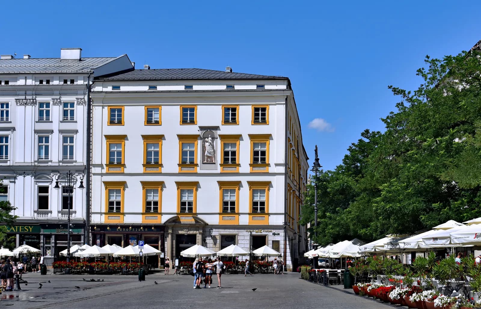 A large white building with yellow-framed windows stands prominently in a city square, with outdoor cafes and people visible below.