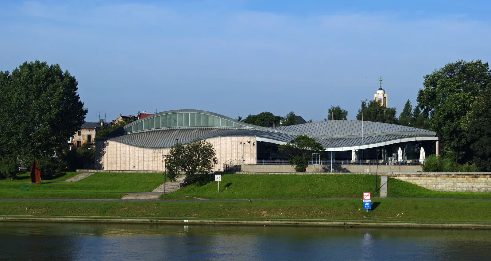 A modern building with a curved roof, light-colored stone walls, and large glass windows overlooks a body of water, with a green lawn and trees in the foreground.